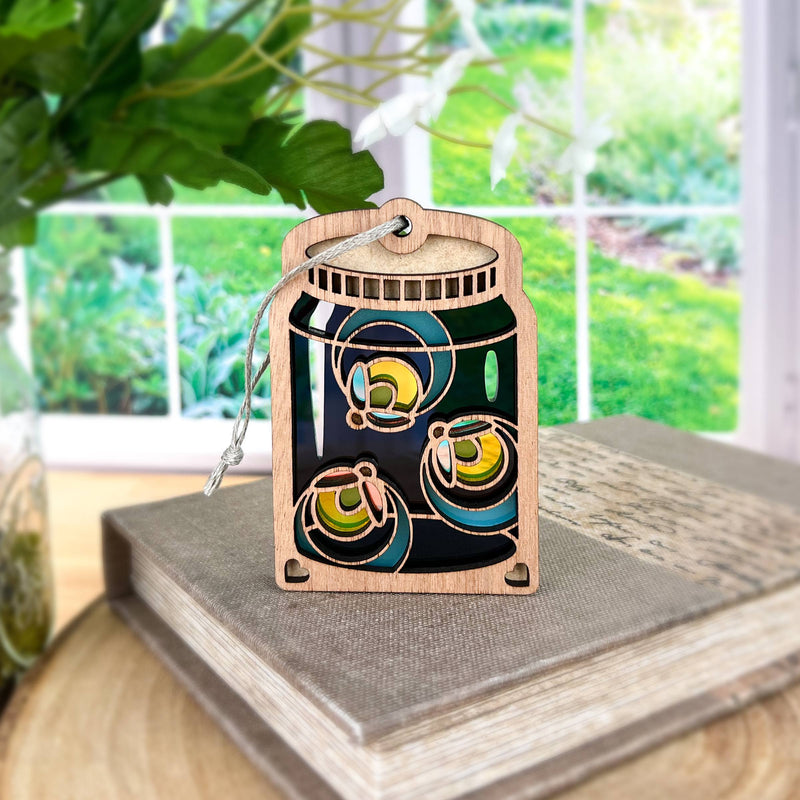 A Forged Flare® Fireflies Jar Ornament, 3.6" tall, sits atop a closed book near a window with a view of a lush green garden.
