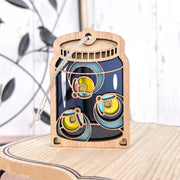 The Forged Flare® Fireflies Jar Ornament, 3.6" (H), features cutout designs of glowing fireflies in a wooden mason jar and hangs by a string, adding a whimsical twist to holiday décor. Displayed on a light wooden surface with a blurred background.