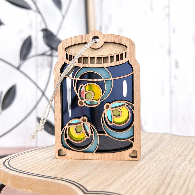 The Forged Flare® Fireflies Jar Ornament, 3.6" (H), features cutout designs of glowing fireflies in a wooden mason jar and hangs by a string, adding a whimsical twist to holiday décor. Displayed on a light wooden surface with a blurred background.