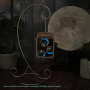 The Forged Flare® Fireflies Jar Ornament, 3.6" (H), glows in the dark with three colorful fireflies. It hangs from a metal stand, with a glowing moon and potted plants behind. Glow acrylic recharges in light, just like real fireflies!.
