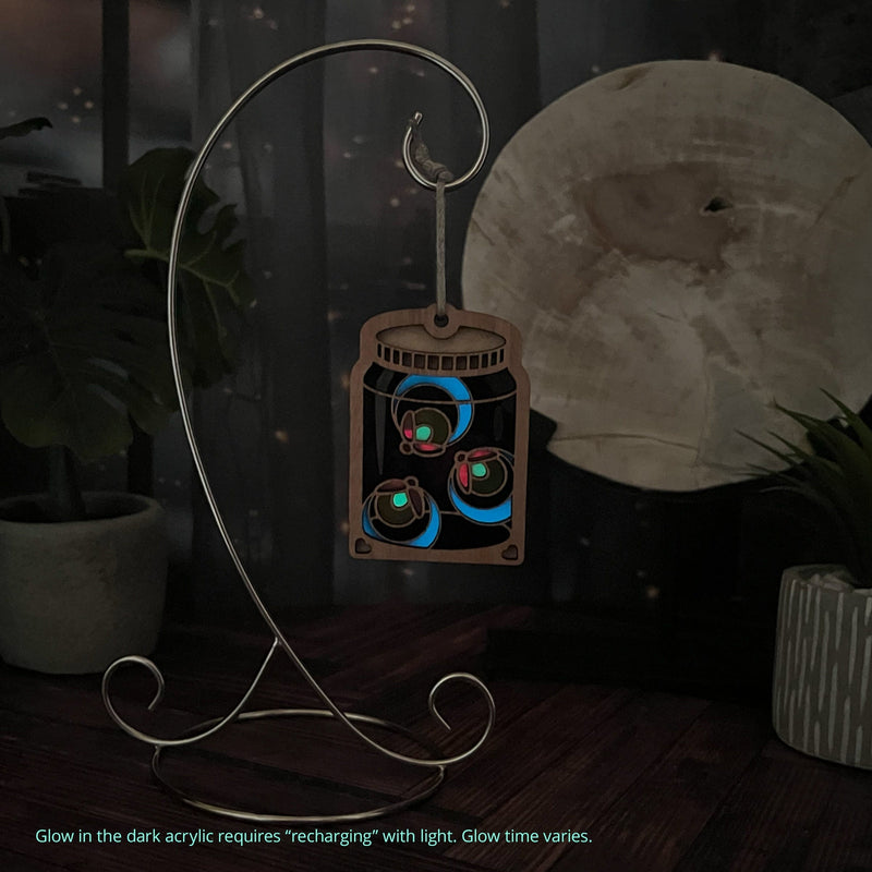 The Forged Flare® Fireflies Jar Ornament, 3.6" (H), glows in the dark with three colorful fireflies. It hangs from a metal stand, with a glowing moon and potted plants behind. Glow acrylic recharges in light, just like real fireflies!.