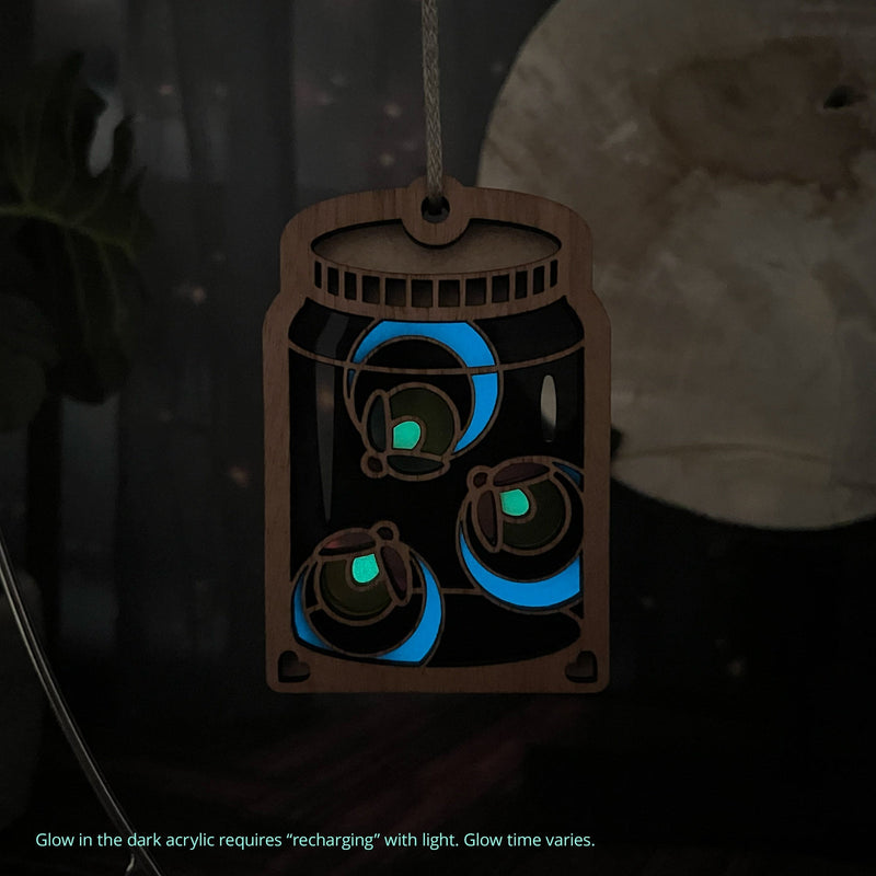 The Forged Flare® Fireflies Jar Ornament, 3.6" (H), features glowing blue and green acrylic fireflies in a wooden mason jar. Glow time varies and the ornament requires "recharging" with light for its magical effect.