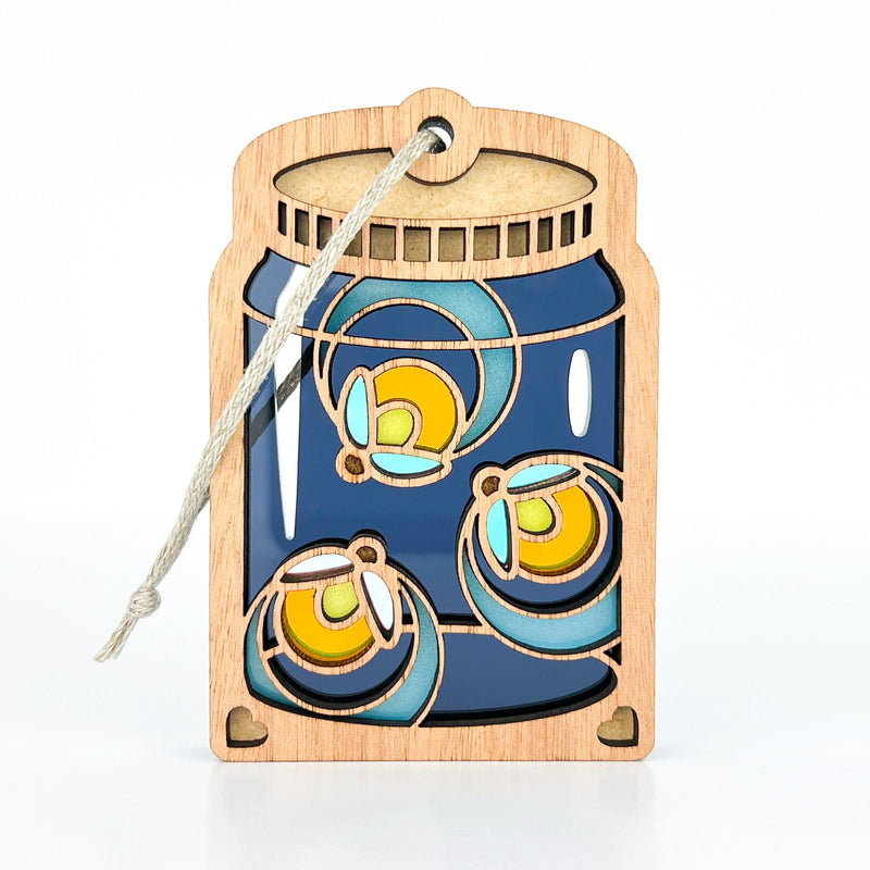 The Forged Flare® Fireflies Jar Ornament, 3.6" (H), features a layered blue, yellow, and teal wood design resembling fireflies in a mason jar with a gray hanging string threaded through the top hole for easy display.