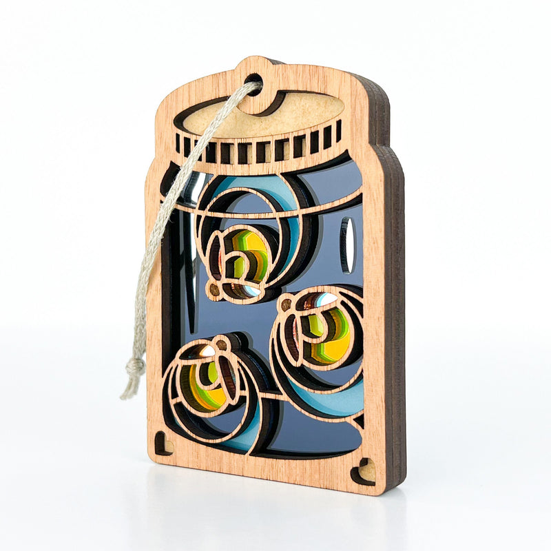 The Forged Flare® Fireflies Jar Ornament, 3.6" (H), features layered wooden cutouts of three fireflies with colorful wings inside a mason jar shape, and includes an attached hanging string. The background is plain white.
