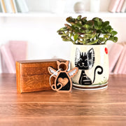 A potted plant in a ceramic cat pot, a wooden box, and the Forged Flare® Forever Loved Cat Angel Ornament for Charity sit on a wooden table. Shelves with books and decor are softly blurred in the background.