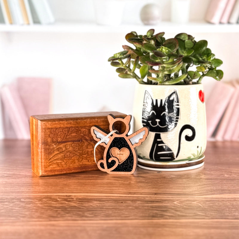 A potted plant in a ceramic cat pot, a wooden box, and the Forged Flare® Forever Loved Cat Angel Ornament for Charity sit on a wooden table. Shelves with books and decor are softly blurred in the background.