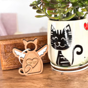A Forged Flare® Forever Loved Cat Angel Ornament for Charity stands beside a ceramic pot featuring a smiling black cat and a green succulent, with a carved wooden box in the background.