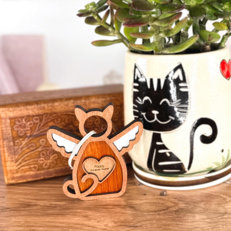 The Forged Flare® Forever Loved Cat Angel Ornament for Charity features a wooden cat figurine with angel wings and a heart engraving, placed before a box and a smiling black cat pot—perfect as a personalized, heartfelt cat memorial.