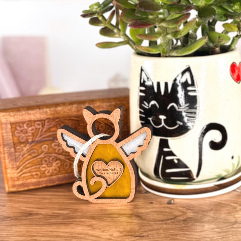 The Forever Loved Cat Angel Ornament for Charity by Forged Flare®—a wooden cat angel with white wings—stands before a smiling black cat planter; to the left, a floral-carved wooden box rests on the surface.