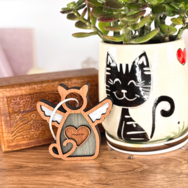 The Forged Flare® Forever Loved Cat Angel Ornament for Charity is a wooden angel cat engraved with "Meegoran," shown beside a succulent in a pot with a black cat and heart, with an ornate box ideal for a cat memorial display.