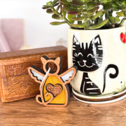 The Forever Loved Cat Angel Ornament for Charity by Forged Flare®—a winged wooden cat with heart—stands before a plant pot with a smiling black cat; a carved wooden box is in the background.
