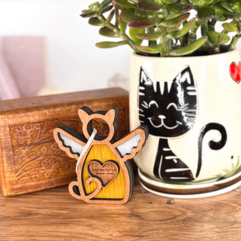 The Forever Loved Cat Angel Ornament for Charity by Forged Flare®—a winged wooden cat with heart—stands before a plant pot with a smiling black cat; a carved wooden box is in the background.
