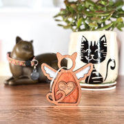 The Forged Flare® Forever Loved Cat Angel Ornament for Charity is a wooden figurine with wings and a heart, engraved with "Korhu Street 2004-2018." Behind it sit a cat-shaped statue and a plant in a pot with a cat face design.