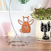 A Forged Flare® Forever Loved Cat Angel Ornament for Charity hangs on a metal stand, while a custom cat planter with succulent sits nearby. Books and decor in the background add extra charm.