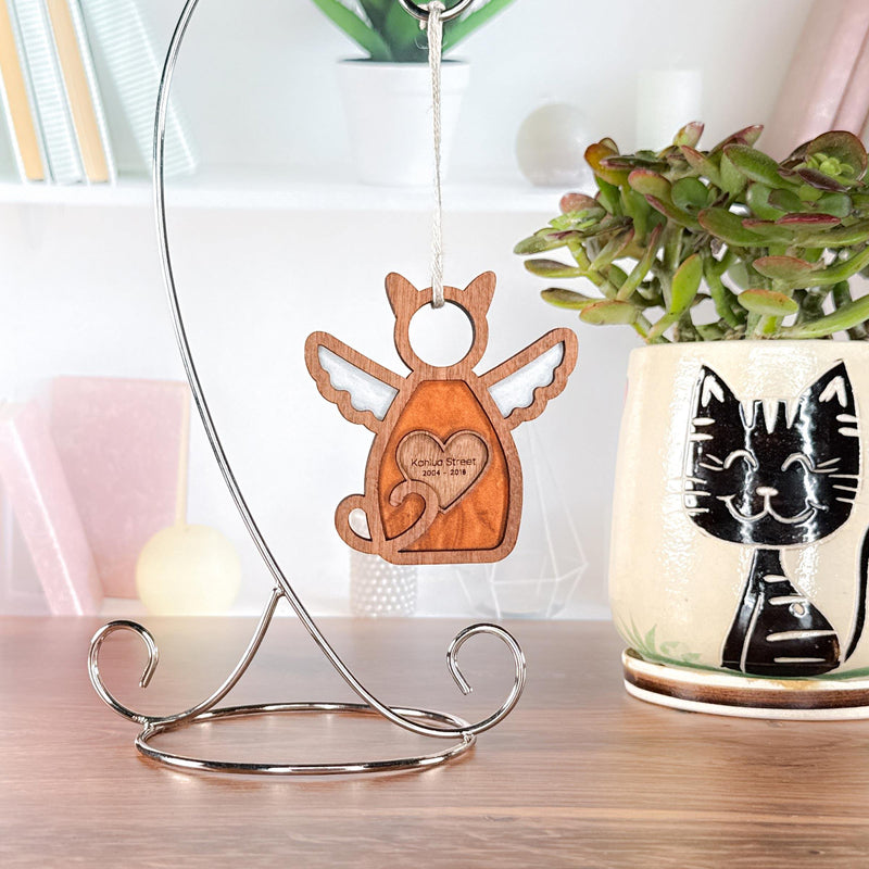 A Forged Flare® Forever Loved Cat Angel Ornament for Charity hangs on a metal stand, while a custom cat planter with succulent sits nearby. Books and decor in the background add extra charm.