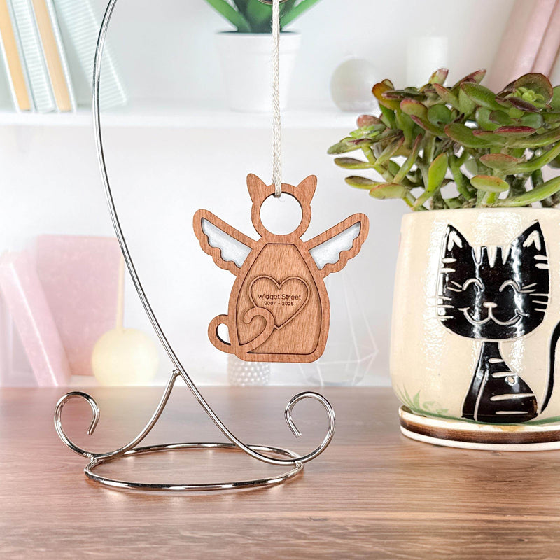 The Forged Flare® Forever Loved Cat Angel Ornament for Charity hangs from a silver stand on a desk, accompanied by a succulent in a black cat vase and books, creating a charming display.