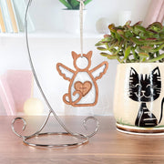 The Forged Flare® Forever Loved Cat Angel Ornament for Charity features a wooden angel-cat with wings and a heart on a silver stand. Behind it, a mug with a black cat design holds a plant, next to books on white shelves.