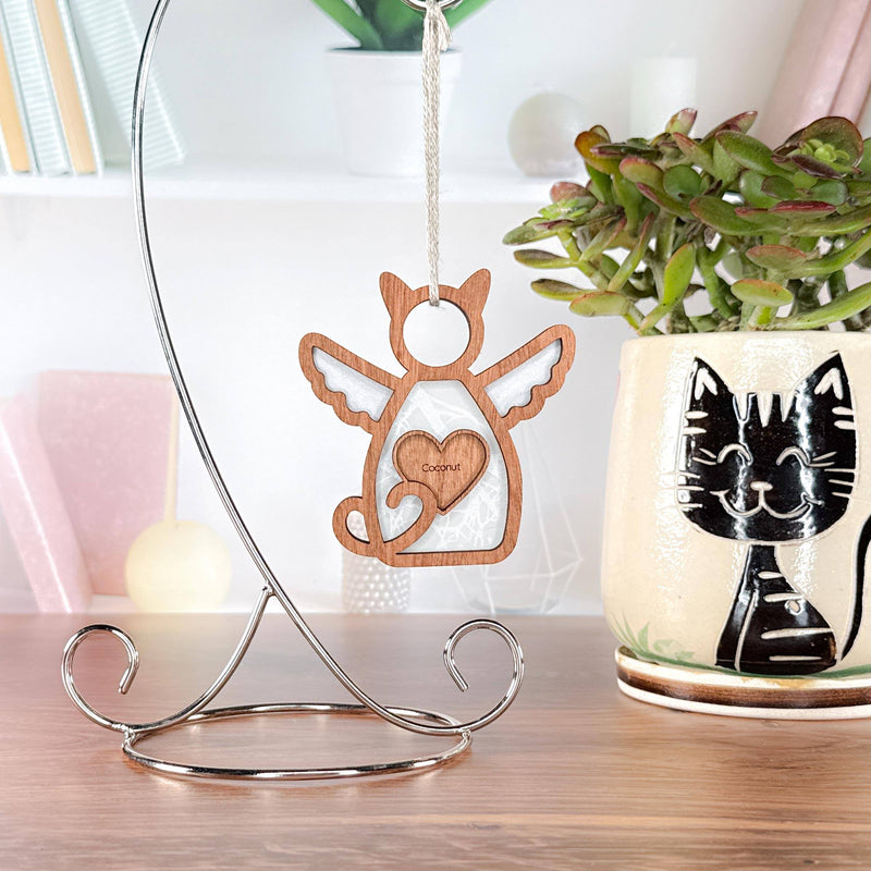 The Forged Flare® Forever Loved Cat Angel Ornament for Charity features a wooden angel-cat with wings and a heart on a silver stand. Behind it, a mug with a black cat design holds a plant, next to books on white shelves.
