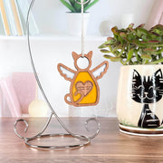 The Forged Flare® Forever Loved Cat Angel Ornament for Charity hangs on a metal stand beside a cat mug with a plant, set on a desk—an ideal, heartfelt addition to any cat memorial or tribute space.