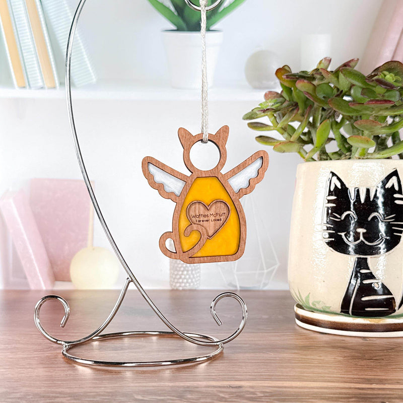 The Forged Flare® Forever Loved Cat Angel Ornament for Charity hangs on a metal stand beside a cat mug with a plant, set on a desk—an ideal, heartfelt addition to any cat memorial or tribute space.