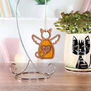 The Forged Flare® Forever Loved Cat Angel Ornament for Charity hangs from a metal stand on a desk. A plant in a mug with a black cat design sits nearby. The ornament, with wings and tail, makes a touching cat memorial.
