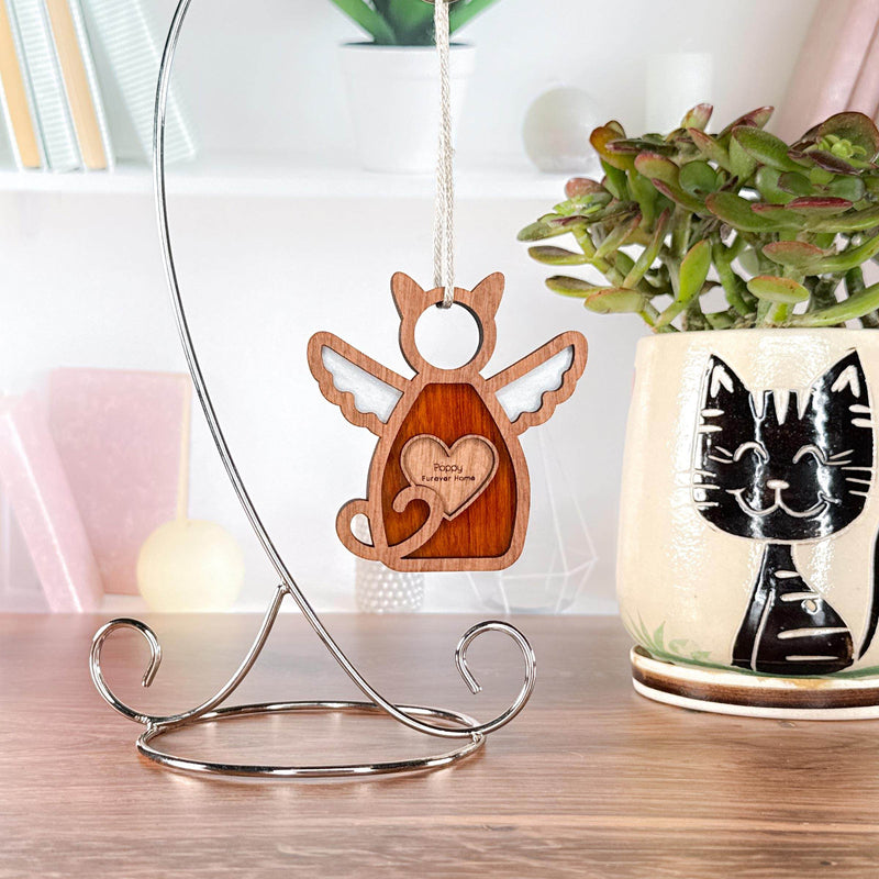 The Forged Flare® Forever Loved Cat Angel Ornament for Charity, featuring a heart cutout, hangs from a stand as a cat memorial. Nearby sits a black cat face ceramic planter with green succulent, set among books and decor.