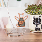 The Forged Flare® Forever Loved Cat Angel Ornament for Charity hangs from a silver stand on a desk beside a potted succulent in a cat-face mug, with shelves and books behind—creating a touching cat memorial display.