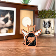 The Forged Flare® Forever Loved Cat Angel Ornament for Charity is a wooden cat-shaped angel with wings and a heart, engraved with "Gizmo," perfect as a cat memorial, shown beside a framed photo, white paw print mold, and illustrated cat mug.