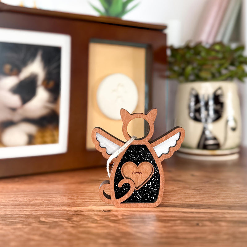 The Forged Flare® Forever Loved Cat Angel Ornament for Charity is a wooden cat-shaped angel with wings and a heart, engraved with "Gizmo," perfect as a cat memorial, shown beside a framed photo, white paw print mold, and illustrated cat mug.