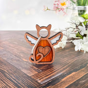 The Forged Flare® Forever Loved Cat Angel Ornament for Charity is a wooden angel-shaped ornament with wings, a heart cutout, and heart design—ideal as a custom cat ornament—shown on wood with white and pink flowers behind.