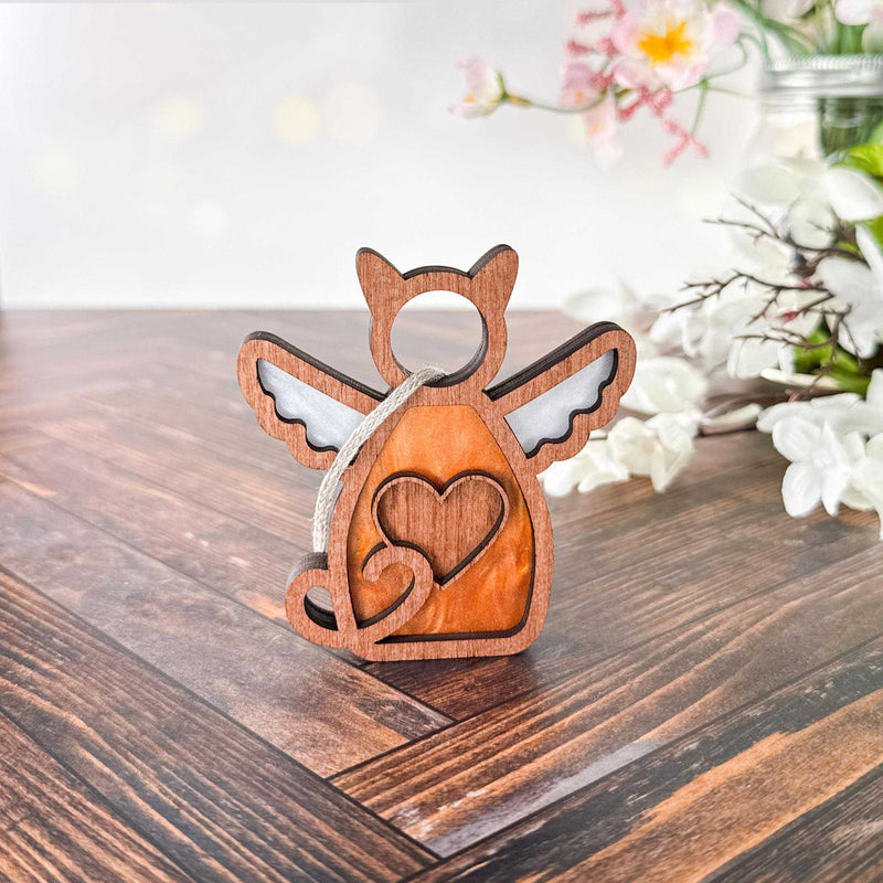 The Forged Flare® Forever Loved Cat Angel Ornament for Charity, a wooden cat-shaped angel with wings and a heart cutout, stands on a table with soft-focus white flowers in the background.