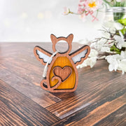 The Forged Flare® Forever Loved Cat Angel Ornament for Charity, a wooden keychain shaped like an angel cat with wings and a heart, rests on wood with blurred white and pink flowers in the background.