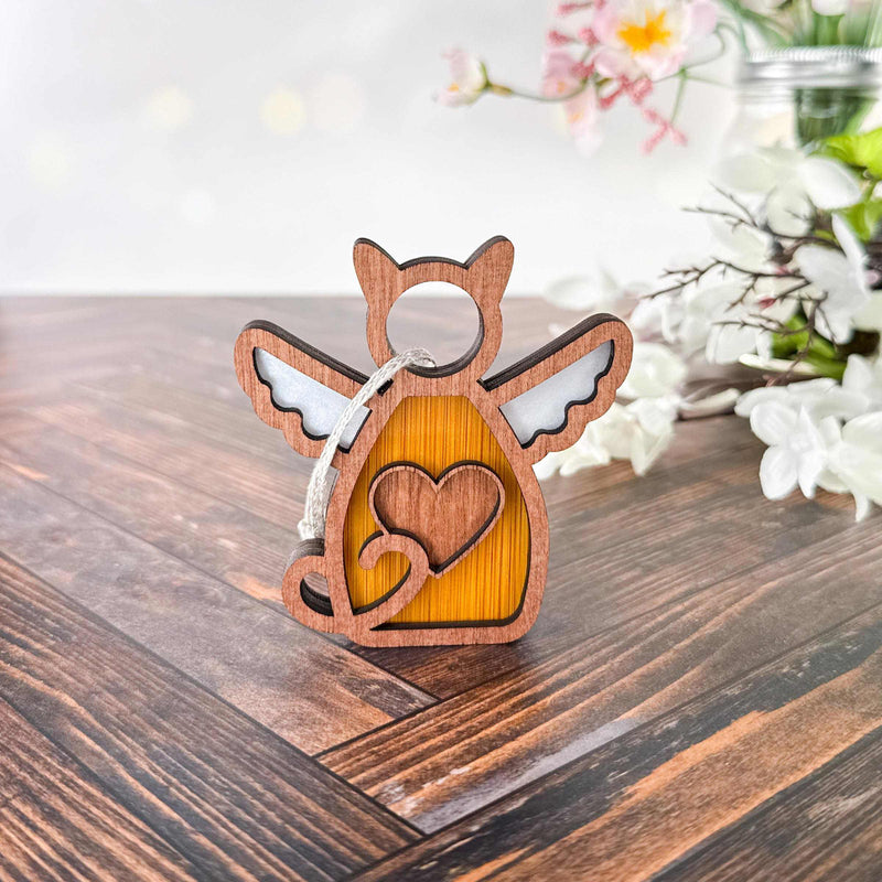 The Forged Flare® Forever Loved Cat Angel Ornament for Charity, a wooden keychain shaped like an angel cat with wings and a heart, rests on wood with blurred white and pink flowers in the background.