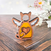The Forged Flare® Forever Loved Cat Angel Ornament for Charity, shaped like a winged cat with a heart, sits on a wooden surface—ideal as a personalized pet ornament or cat memorial. Blurred white and pink flowers appear in the background.