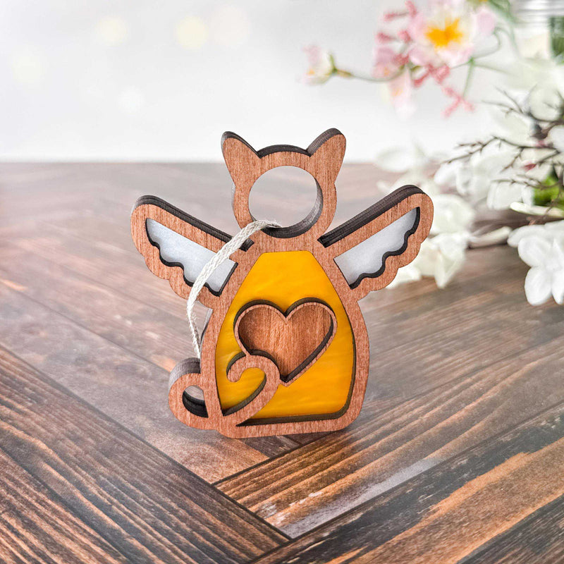 The Forged Flare® Forever Loved Cat Angel Ornament for Charity, shaped like a winged cat with a heart, sits on a wooden surface—ideal as a personalized pet ornament or cat memorial. Blurred white and pink flowers appear in the background.