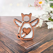 The Forged Flare® Forever Loved Cat Angel Ornament for Charity, shaped like an angel with cat ears, wings, and a heart cutout, rests on a wooden surface with blurred white flowers and green leaves in the background.