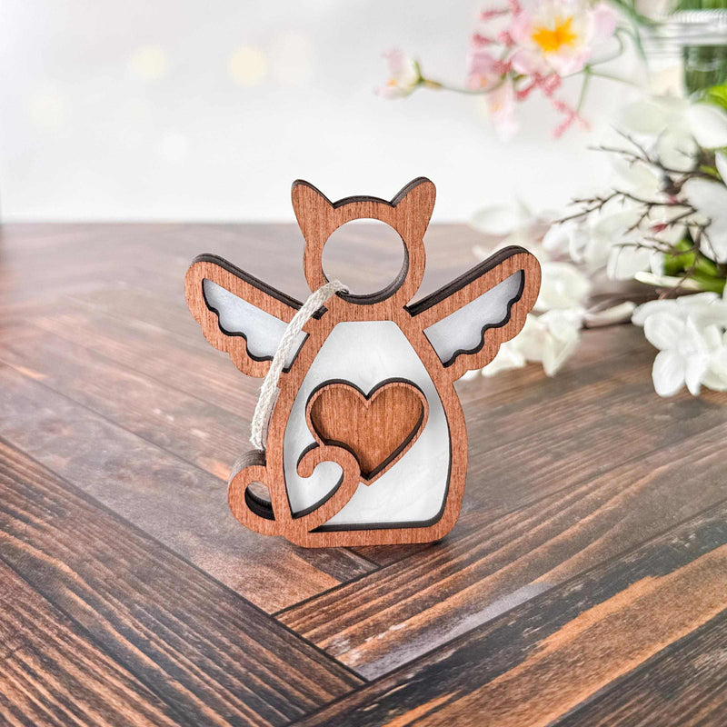 The Forged Flare® Forever Loved Cat Angel Ornament for Charity, shaped like an angel with cat ears, wings, and a heart cutout, rests on a wooden surface with blurred white flowers and green leaves in the background.