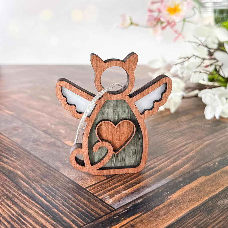 The Forged Flare® Forever Loved Cat Angel Ornament for Charity features a wooden angel with a heart cutout and white wings, standing on wood—perfect for cherishing your pet’s memory. White flowers and greenery are softly blurred in the background.