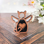 The Forged Flare® Forever Loved Cat Angel Ornament for Charity sits on a wooden surface, featuring angel wings and a heart cutout, with white flowers and greenery softly blurred in the background.