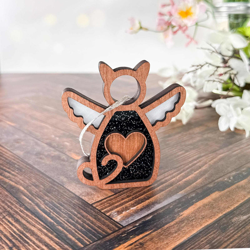 The Forged Flare® Forever Loved Cat Angel Ornament for Charity sits on a wooden surface, featuring angel wings and a heart cutout, with white flowers and greenery softly blurred in the background.
