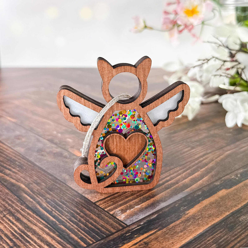 The Forged Flare® Forever Loved Cat Angel Ornament for Charity features a winged cat with a heart cutout, filled with colorful confetti, hanging by a string on wood with white flowers—a touching personalized pet memorial.