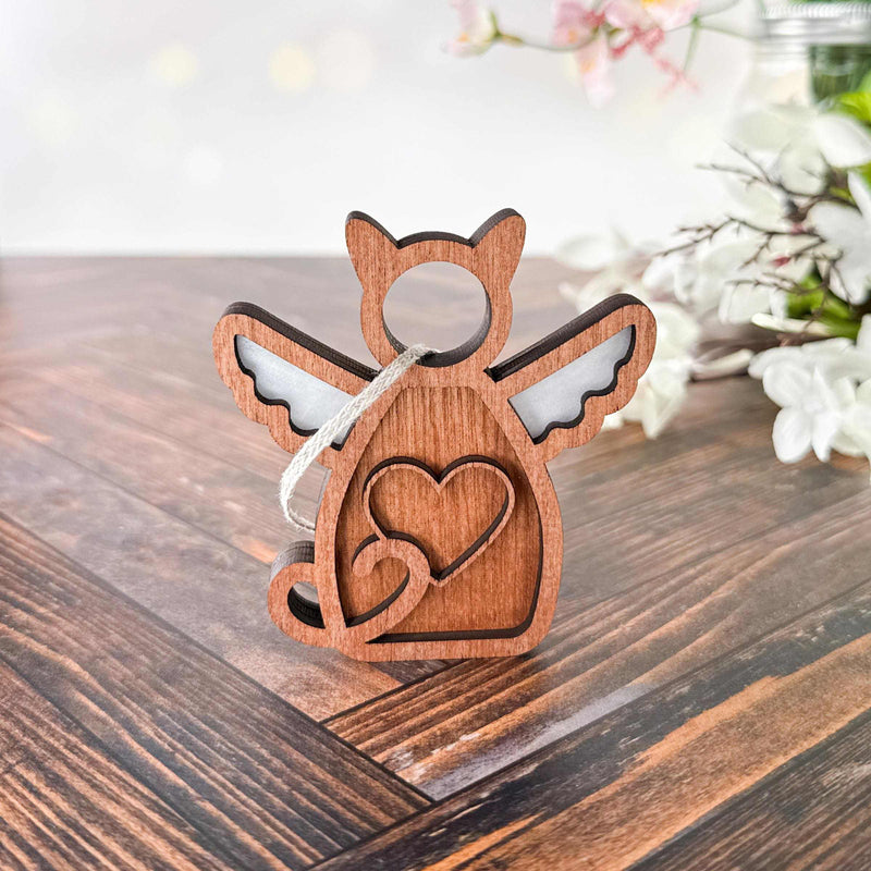 The Forged Flare® Forever Loved Cat Angel Ornament for Charity, featuring angel wings and a heart cutout, stands on a wooden surface with blurred white flowers in the background.
