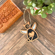 The Forged Flare® Forever Loved Cat Angel Ornament for Charity—a wooden angel-shaped keepsake with white wings, heart detail, and black glitter accents—rests by a carved box and potted plant, honoring cherished cat memories.