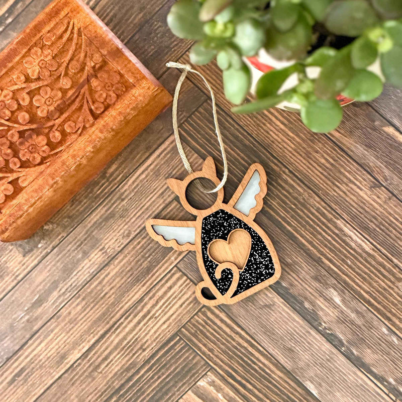 The Forged Flare® Forever Loved Cat Angel Ornament for Charity—a wooden angel-shaped keepsake with white wings, heart detail, and black glitter accents—rests by a carved box and potted plant, honoring cherished cat memories.