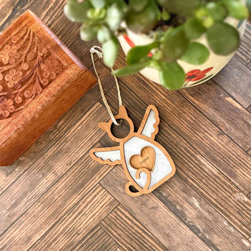 The Forged Flare® Forever Loved Cat Angel Ornament for Charity, featuring cut-out wings and a curled tail, rests on a wooden floor by a carved box and potted plant—an ideal personalized cat memorial keepsake.