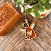 The Forged Flare® Forever Loved Cat Angel Ornament for Charity—a wooden cat-shaped memorial with angel wings and heart, inscribed “Poco’s Momma”—rests by a carved box and plant, offering a heartfelt, personalized tribute.