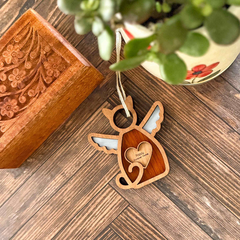The Forged Flare® Forever Loved Cat Angel Ornament for Charity—a wooden cat-shaped memorial with angel wings and heart, inscribed “Poco’s Momma”—rests by a carved box and plant, offering a heartfelt, personalized tribute.