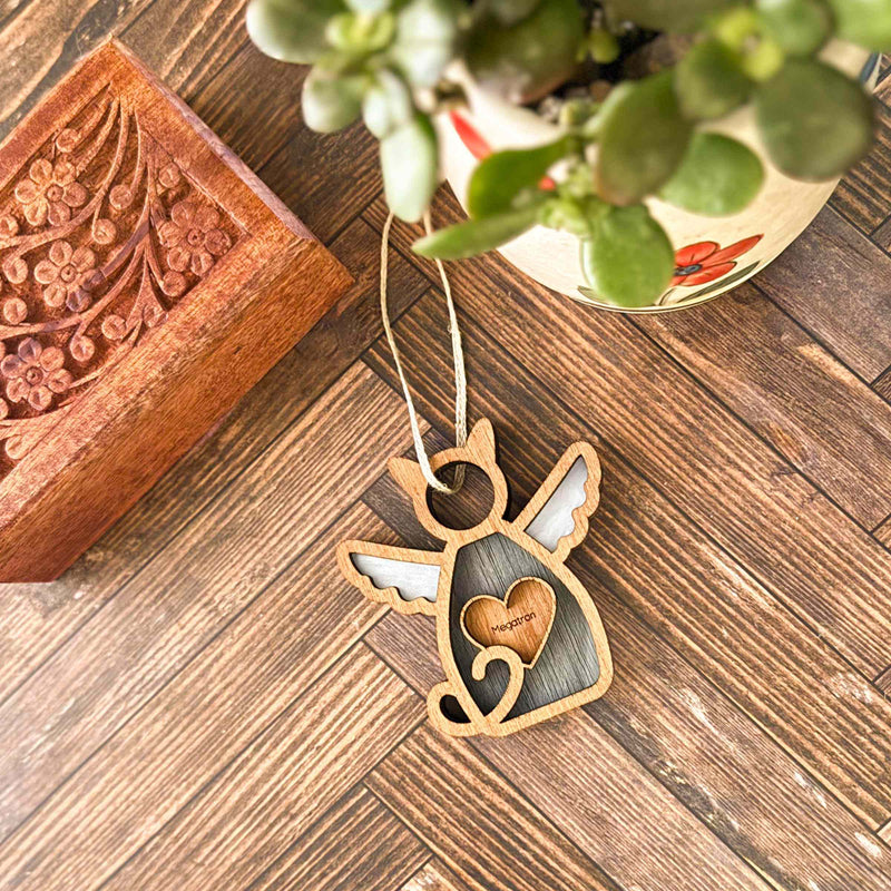 A Forged Flare® Forever Loved Cat Angel Ornament for Charity lies on a wooden floor next to a carved box, potted succulent, and engraved "Mommy" angel-shaped ornament.