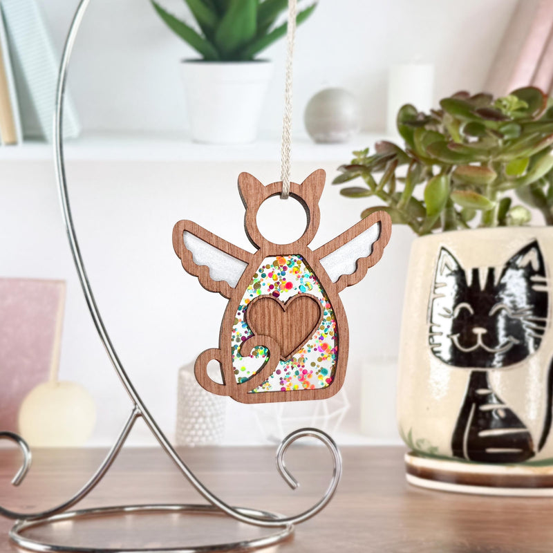 The Forged Flare® Forever Loved Cat Angel Ornament for Charity, featuring wings and a heart cutout, hangs on a metal stand near a potted plant and a ceramic mug with a black cat face, all arranged on a wooden surface.