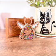 The Forged Flare® Forever Loved Cat Angel Ornament for Charity is a wooden, cat-shaped figure with wings, filled with colorful beads and labeled "Cinnamon My Sweet Spice," displayed near a plant in a cat mug and a wooden box.